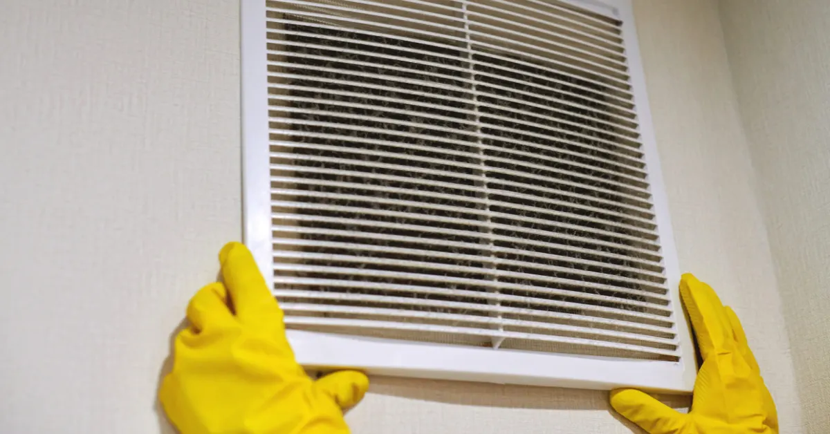 Gloved hands removing a dust-covered return air vent cover from a wall
